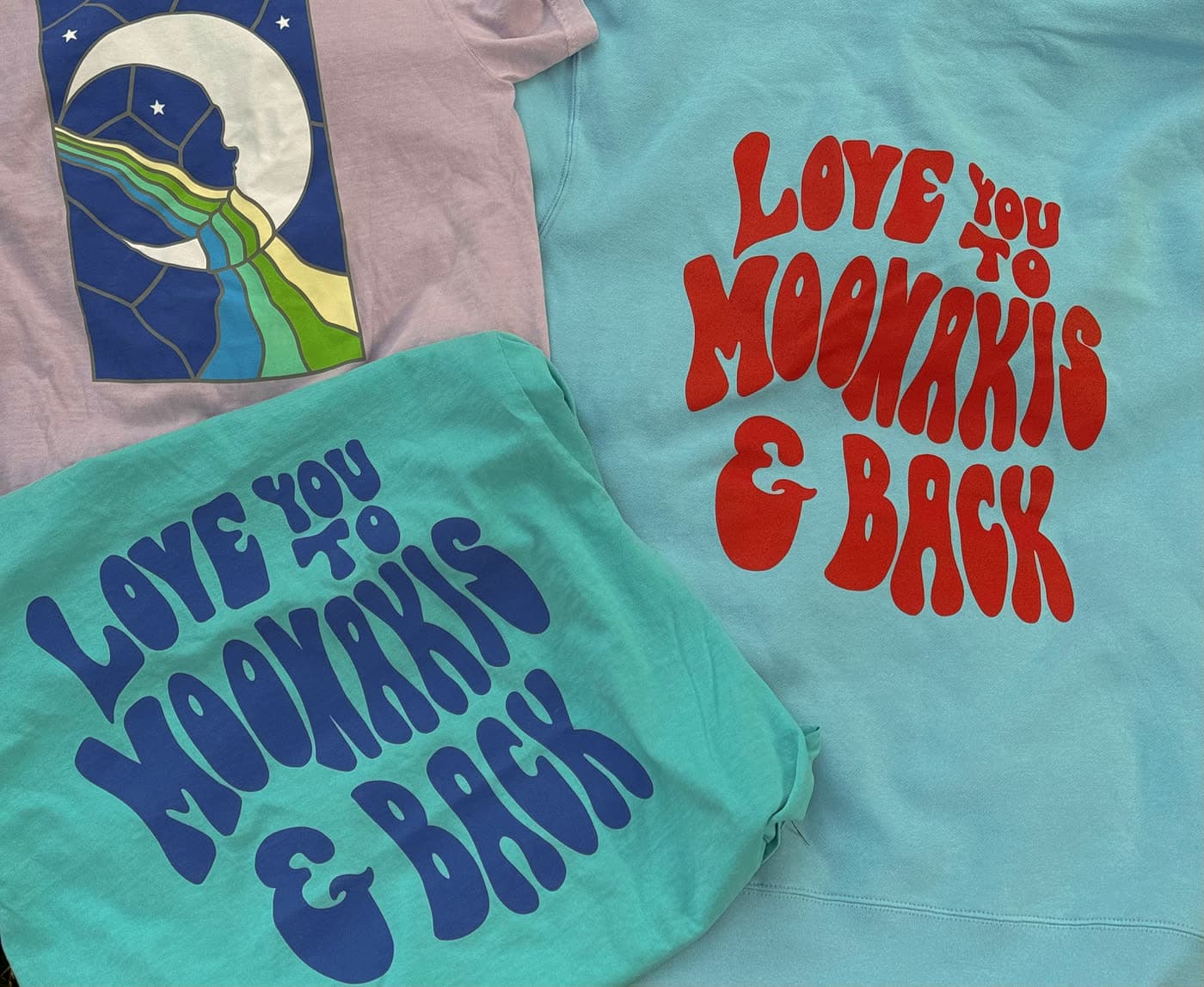 Three shirts laid flat: a lavender tee with a stained-glass moon print, and teal and light blue shirts reading Love you to Moonakis and back