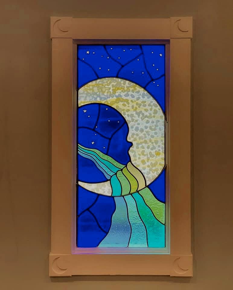A stained-glass window of a crescent moon with a face, a river of green and blue glass flowing from it, set in a moon-carved wooden frame