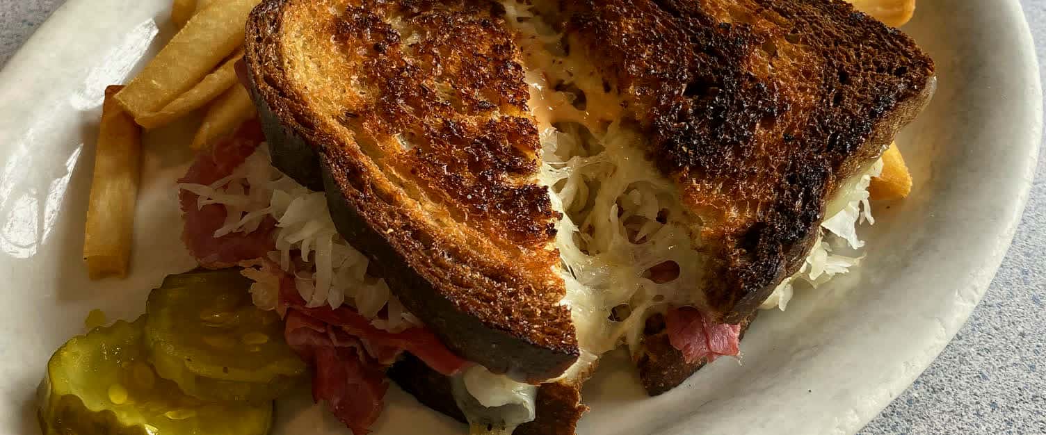A grilled Reuben cut in halves with fries and pickles on a diner plate