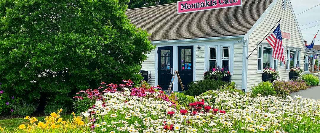 The white Moonakis Cafe building with its rooftop sign and an American flag, a summer flower garden in front