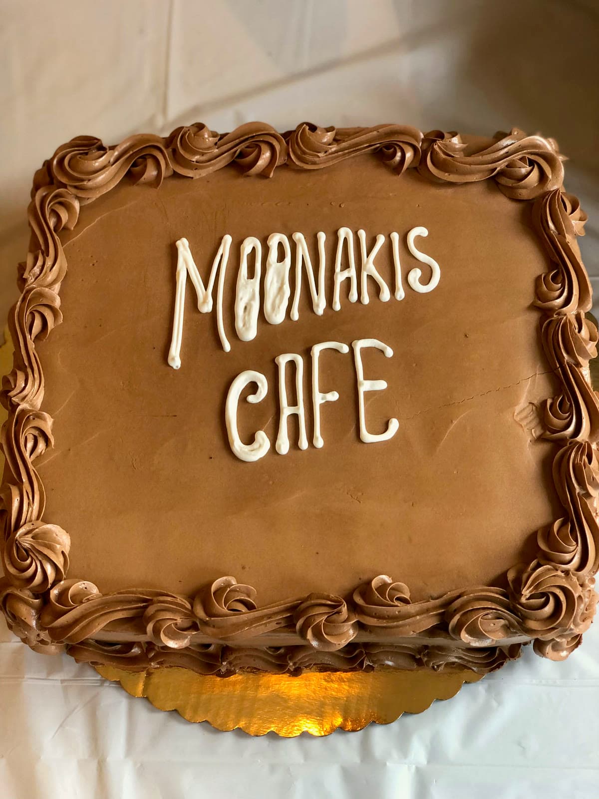 Square chocolate cake with a piped chocolate buttercream border and MOONAKIS CAFE written in white icing, two moons dotting the O's
