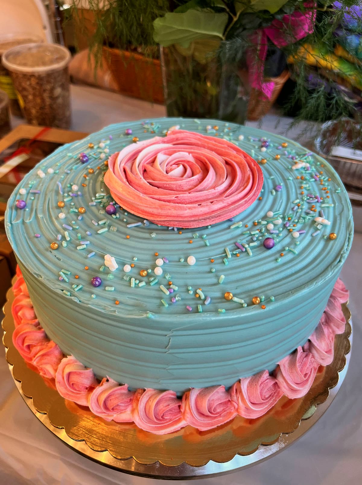 Round cake in combed sea-glass teal buttercream with a large pink rosette on top, sprinkles, and a pink rosette border on a gold board