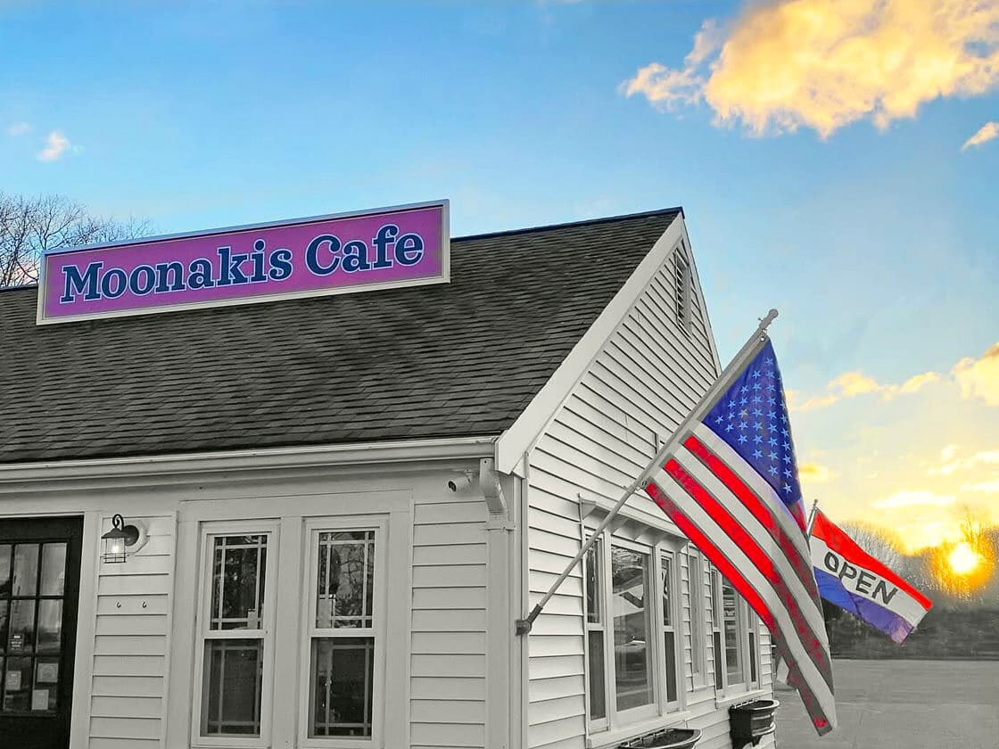 The corner of the Moonakis Cafe building at golden hour, its rooftop sign lit, with an American flag and a glowing OPEN sign