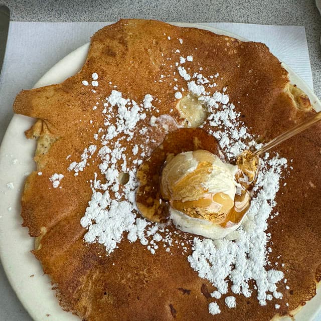 Maple syrup pouring onto a plate-wide griddle pancake with whipped butter and powdered sugar